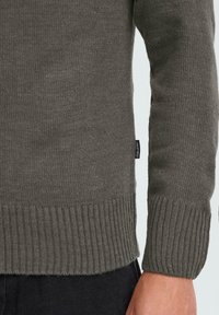 Dunkelgrüner, gestrickter Pullover mit gerippten Bündchen und Saum. Enthält ein kleines Markenetikett an der Seite. Strukturierter Wollmischstoff.