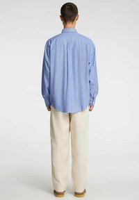 Gaiši zila un balta svītraina krekliņa ar pogu apkakli, brīva piegriezuma un apaļu apmali, kopā ar krēmkrāsas biksēm.