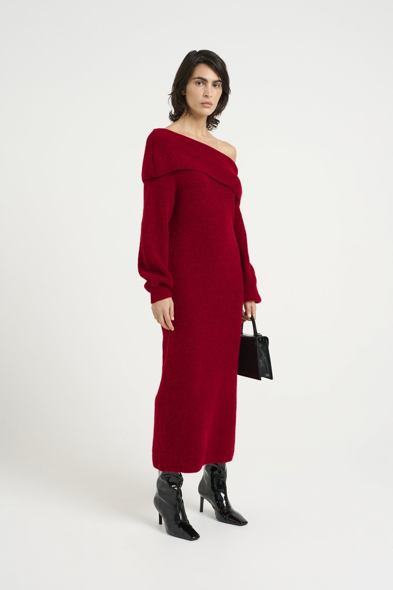 Ein langes rotes schulterfreies Kleid mit langen Ärmeln und einem strukturierten Strickmuster, kombiniert mit schwarzen glänzenden Ankle Boots und einer kleinen schwarzen Handtasche.