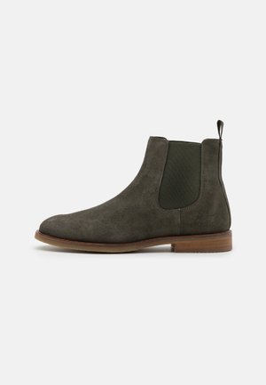 Bota Chelsea de ante color oliva oscuro con panel lateral elástico, lengüeta para tirar y tacón bajo apilado marrón sobre fondo blanco.