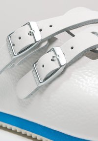 White leather sandal with two adjustable silver buckles, textured upper, and a blue and white sole. Features cutouts for ventilation.
