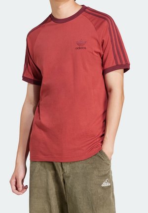 Homme portant un t-shirt rouge Adidas avec trois bandes sur les manches et un pantalon en velours côtelé vert olive Adidas, une main dans la poche.