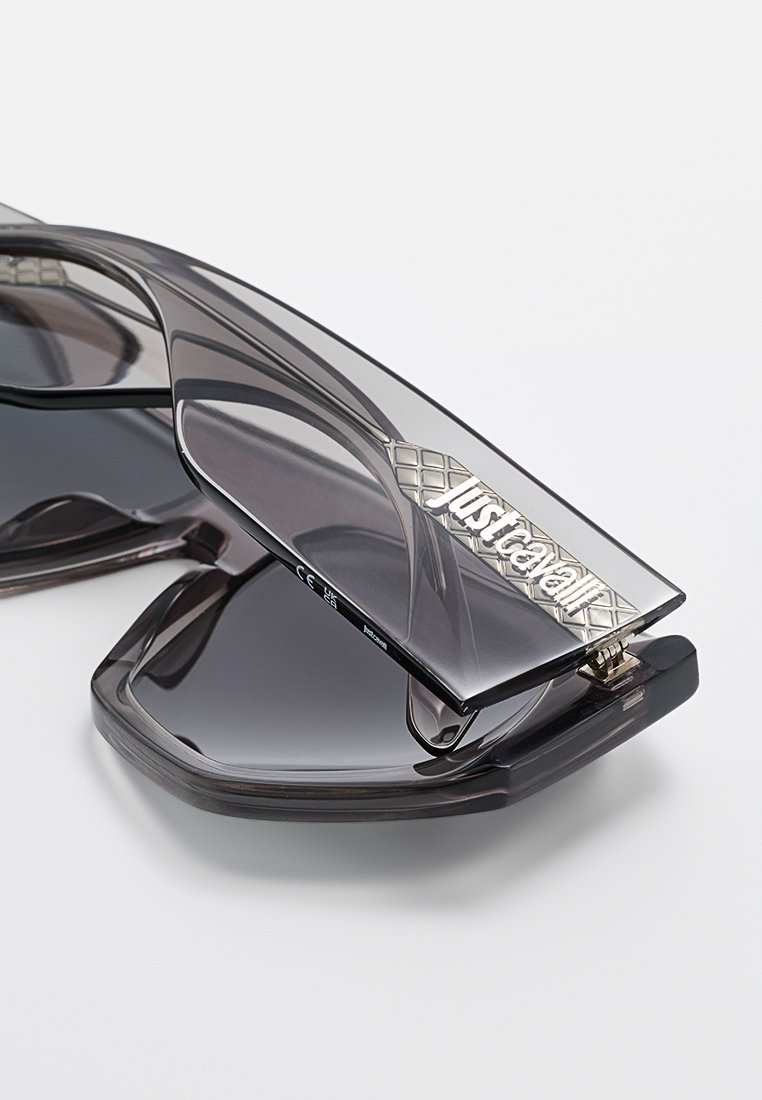 Lunettes de soleil avec une monture translucide gris foncé et des verres teintés. Présente un logo en argent texturé sur la branche et des motifs géométriques.