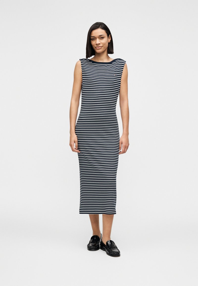 Φόρεμα με ρίγες από ελαστικό ύφασμα· διαθέτει καραβότρατα και αμάνικο σχέδιο. Το φόρεμα φτάνει μέχρι τους αστραγάλους και συνδυάζεται με μαύρα loafers.