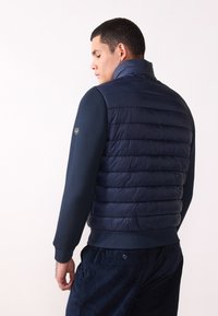 Marinblå quiltad puffer-väst med hög krage, slät textur och figurnära design. Kombinerad med en mörkblå långärmad skjorta.