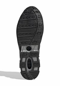 Suela de zapato deportivo negro con goma texturizada, bandas de rodadura visibles, surcos y un elemento de diseño circular en el centro. Presenta detalles plateados.