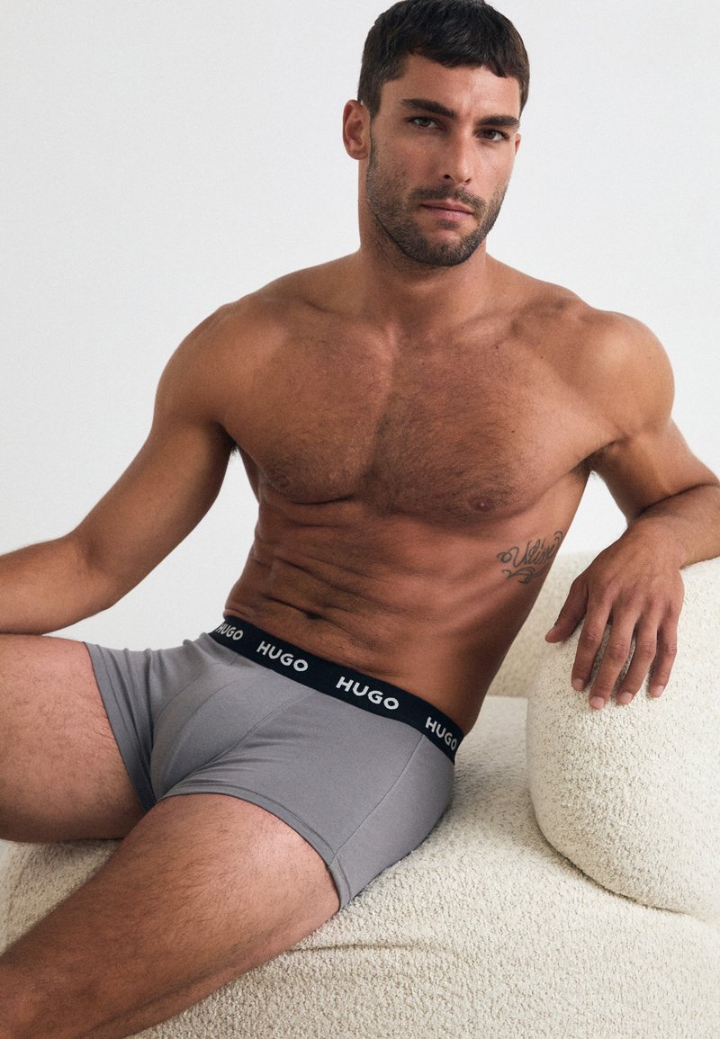 Boxer in jersey grigio con cintura elastica nera etichettata "HUGO," indossati da un modello seduto su una superficie testurizzata di colore crema.