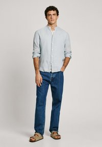 Svetlo modra, dolga sleeva srajca iz lana, oblečena z temno modrimi kavbojkami in rjavimi sandali. Ima sproščen kroj in zasnovo s pasom.