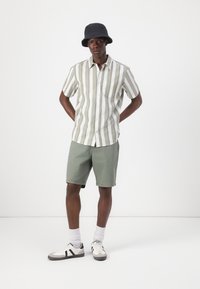 Chemise à manches courtes rayée vert clair et blanche, associée à un short olive vert jusqu'aux genoux. Chapeau seau noir et baskets blanches.