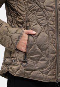 Veste matelassée marron avec une poche zippée, des boutons-pression et une texture lisse. Présente des coutures courbes et un design à coupe décontractée.