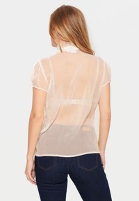 Blouse beige clair et léger avec des manches courtes bouffantes, un col à volants et un motif à pois subtil ; associée à un jean bleu foncé.