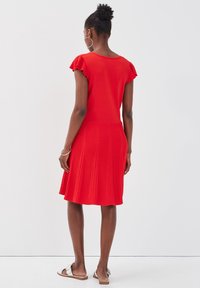 Robe tricotée rouge avec des manches courtes à volants, jupe plissée texturée et corsage uni. Portée avec des sandales plates, vue de dos.