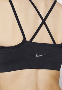 Zwarte sportbeha met gekruiste banden, textuurstof en een zilveren Nike-logo aan de zijkant. Minimalistisch ontwerp en aansluitende pasvorm.