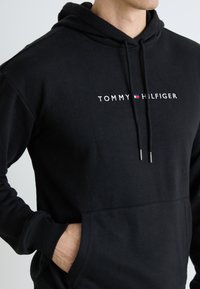 Mann som har på seg en svart Tommy Hilfiger hettegenser med logo sentrert på brystet og hånden i frontlommen, vist fra nakke til midje.
