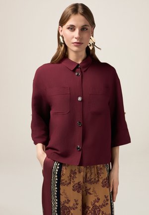 Camisa burdeos de botones con dos bolsillos en el pecho y mangas cortas, combinada con pantalones estampados que presentan acentos florales y rayados.