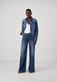 Giacca di jeans con tonalità blu, due tasche sul petto e bottoni argentati, abbinata a jeans blu a vita alta e gamba larga, con orli sfilacciati.