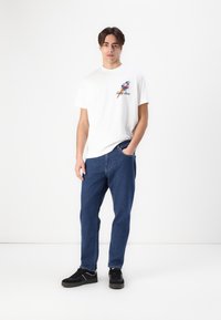 Hvid bomulds-t-shirt med et farverigt fuglemotiv på brystet, parret med blå denimjeans og sorte sneakers. Afslappet design, lige pasform.