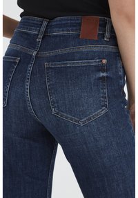 Jeans de denim azul oscuro con cintura alta, que presentan una etiqueta de cuero, costuras dobles y dos bolsillos traseros, mostrados desde un ángulo lateral.