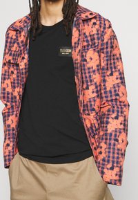 Red and navy plaid jacket with a textured finish over a black T-shirt featuring a gold Moschino logo patch. Tan trousers complete the look.