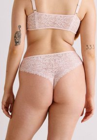 Femme portant un soutien-gorge en dentelle rose clair et des panty assortis, vue de dos, avec des tatouages sur les deux bras.