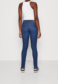 Smala jeans i mörkblå denim med en texturerad yta, med två bakfickor och en brun läderetikett vid midjebandet. Svarta sneakers syns.