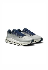 Hellgraue Sportschuhe mit einem strukturierten Mesh-Obermaterial, marineblauen Schnürsenkeln und einer gepolsterten weißen Sohle, die ovale Ausschnitte für Flexibilität aufweist.