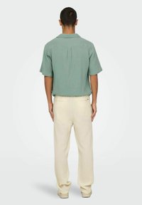 Chemise à manches courtes, de couleur vert clair avec un tissu texturé, associée à un pantalon à pattes large de couleur crème. Détails de design subtils sur les deux vêtements.