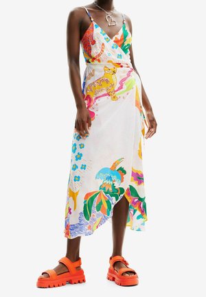 Kobieta ubrana w kolorową sukienkę kopertową z motywami zwierzęcymi i kwiatowymi, zestawioną z masywnymi, jaskrawo pomarańczowymi sandałami oraz naszyjnikiem w kształcie serca.