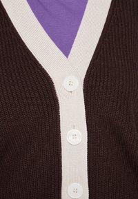 Cardigán marrón con relieve y cuello en V color crema, con cuatro botones redondos. Lleva puesto sobre una camisa morada. Tela de punto texturizada.