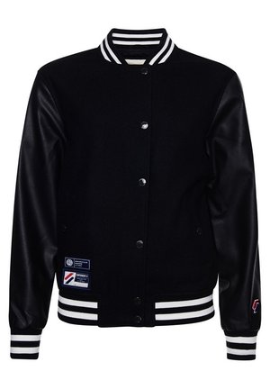 Personne portant une veste universitaire noire, un brassière de sport noire, un pantalon noir évasé et des baskets blanches, debout contre un mur blanc.