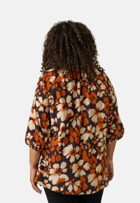 Blusa floreale con maniche corte a palloncino, caratterizzata da modelli astratti arancioni, crema e marroni su uno sfondo scuro. Vestibilità rilassata.