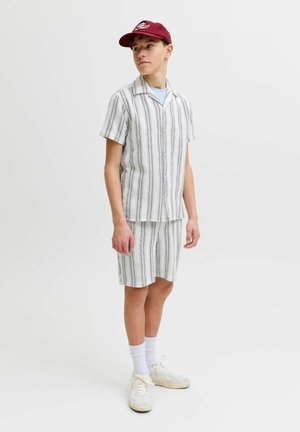 Noor mees, kes kannab valget ja musta triibulist lühikeste varrukatega särki ja sobivaid lühikesi pükse, valgeid tossusid, valgeid sokke ja punast nokamütsi, seisab ja vaatab küljele.