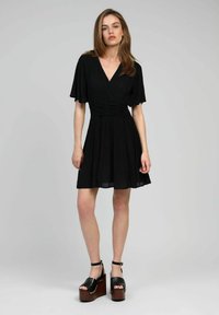 Robe noire courte avec un décolleté en V, des manches flottantes et des détails froncés à la taille ; assortie à des sandales à plateforme noires épaisses.