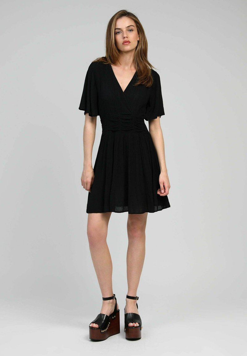 Robe noire courte avec un décolleté en V, des manches flottantes et des détails froncés à la taille ; assortie à des sandales à plateforme noires épaisses.