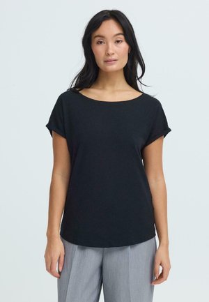 Vrouw met lang donker haar draagt een zwart T-shirt met korte mouwen en lichtgrijze broek, staande tegen een effen lichte achtergrond.