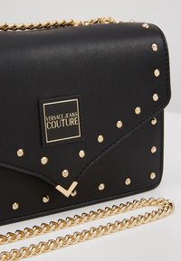 Borsa a tracolla in finta pelle nera con borchie dorate e tracolla a catena. Presenta una placca con logo e una chiusura a pattina con un accento a forma di V.