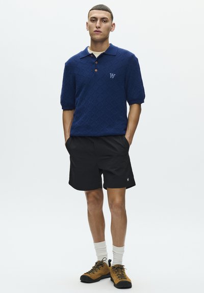Polo bleu marine avec texture en losanges, manches courtes et boutons marron ; associé à un short noir et des chaussures de randonnée marron. Chaussettes blanches visibles.