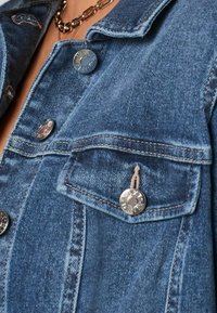 Primer plano del bolsillo y el cuello de una chaqueta de mezclilla azul con botones plateados y collares de cadena dorada en capas visibles en el escote.