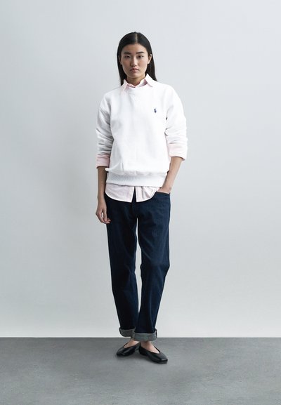 Γυναίκα με μακριά σκούρα μαλλιά στέκεται με το ένα χέρι στην τσέπη, φορώντας λευκό φούτερ πάνω από ανοιχτό ροζ πουκάμισο, σκούρο παντελόνι με μανσέτες και μαύρα μπαλαρίνες.