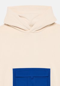 Felpa beige con cappuccio, caratterizzata da una grande tasca frontale blu con un'apposita patta rettangolare. Texture morbida e design semplice.
