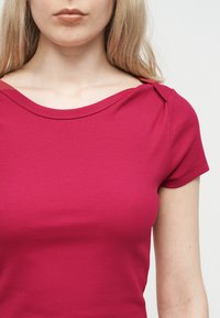 Γυναίκα που φοράει εφαρμοστό, κοντομάνικο, ριμπ πλεκτό τοπ σε ματζέντα χρώμα με φαρδιά λαιμόκοψη, φωτογραφημένη από τους ώμους έως τη μέση μπροστά σε απλό φόντο.