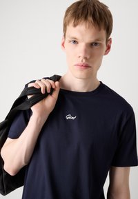 Tricou din bumbac bleumarin, cu guler rotund și mâneci scurte, având un logo brodat alb pe piept. Modelul ține o geantă neagră.