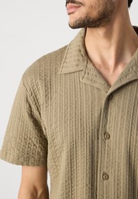 Khaki kortærmet skjorte i struktureret stof med lodrette striber, med en skrå krave og to knapper foran.