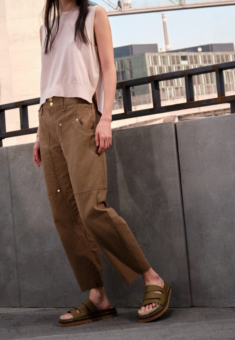 Beige mouwloos topje, bruine utility broek met zakken en gouden accenten, en olijfgroene sandalen met meerdere riemen en een dikke zool.