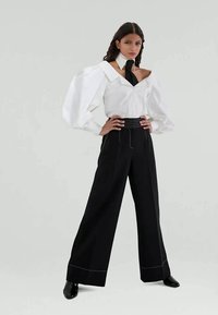 Witte oversized blouse met pofmouwen en zwarte strik, gecombineerd met hoge zwarte broeken met witte stiksels.