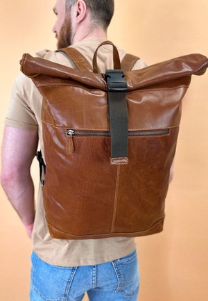 Brown leather roll-top backpack with a smooth texture, adjustable shoulder straps, and a front zip pocket. Subtle stitching details visible.