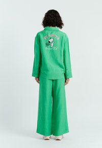 Groen linnen overhemd en wijde broekset, met een Snoopy-afbeelding en de tekst "PEANUTS Racquet Club" op de achterkant. Zachte textuur, casual ontwerp.