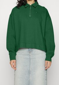 Zelený zip-up pulovr s límcem, vyrobený z texturované látky. Obsahuje malé logo, volný střih a žebrované manžety. Nosen s džínovou sukní.