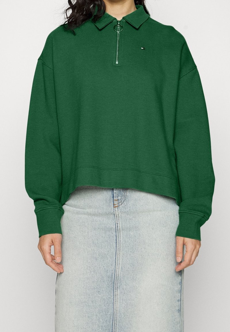 Zelený zip-up pulovr s límcem, vyrobený z texturované látky. Obsahuje malé logo, volný střih a žebrované manžety. Nosen s džínovou sukní.