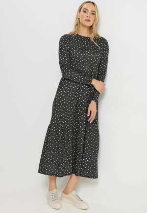 Vrouw staand, gekleed in een zwart midi-jurk met lange mouwen en witte polkadots en beige sneakers, tegen een effen witte achtergrond.